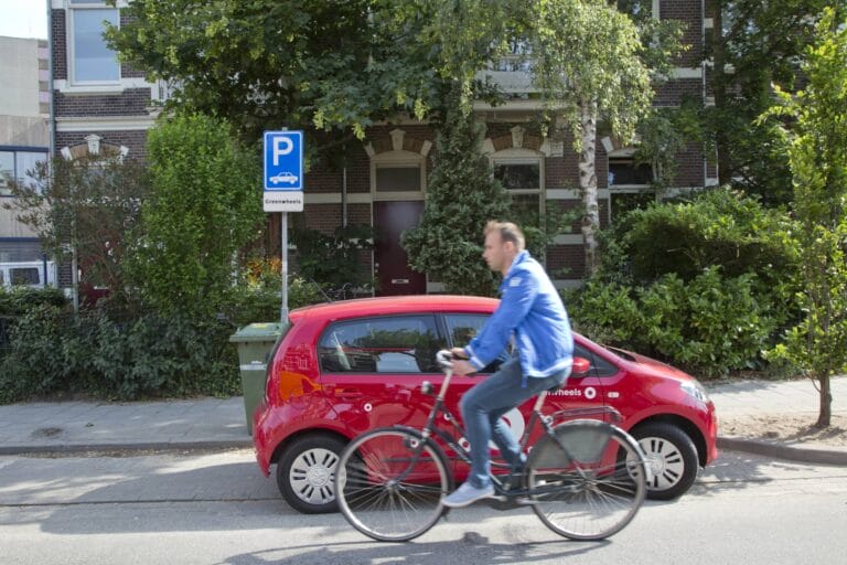 Fietser passeert geparkeerde rode deelauto in straat.